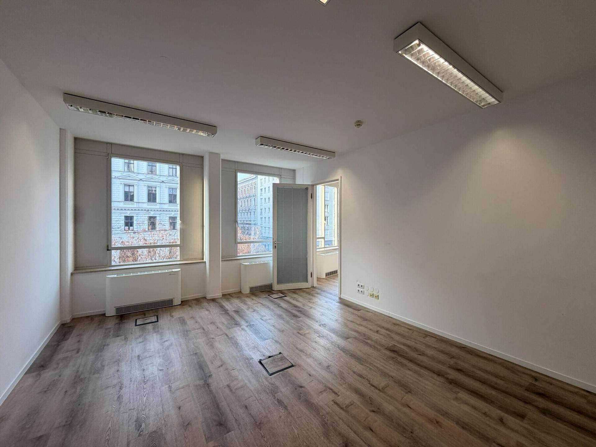 Leerer Büroraum mit Laminatboden und zwei Fenstern, die einen Blick auf die Stadt bieten.