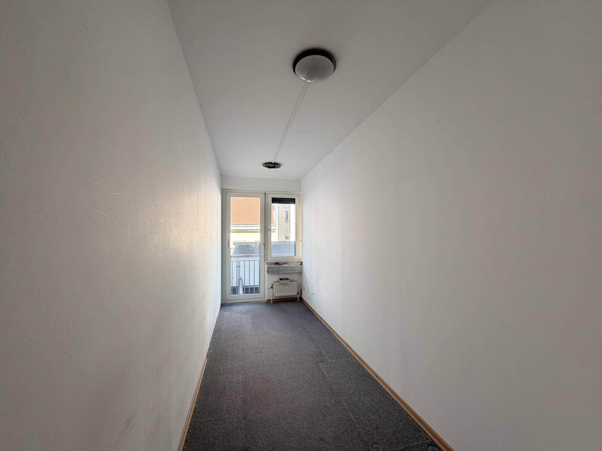 Schmaler Raum mit Teppichboden und einer Tür, die zu einem kleinen Balkon mit Geländer führt.
