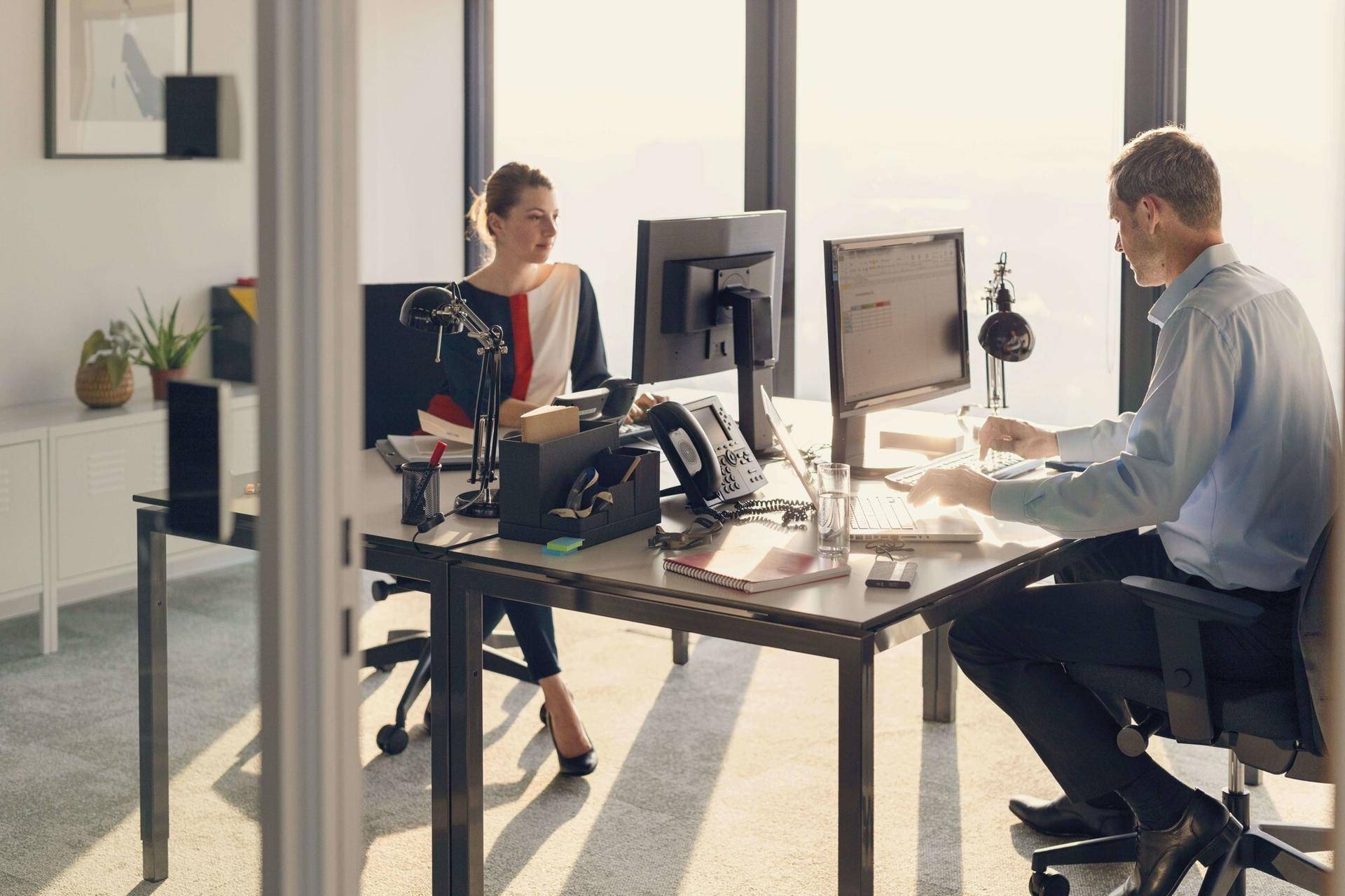 Zwei Personen arbeiten in einem modernen Büro mit großen Fenstern, die viel natürliches Licht hereinlassen.