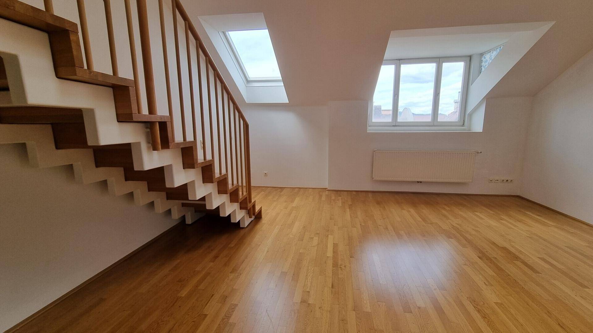 Heller Wohnbereich mit Holztreppe, Dachfenstern und Parkettboden in einem Dachgeschoss.