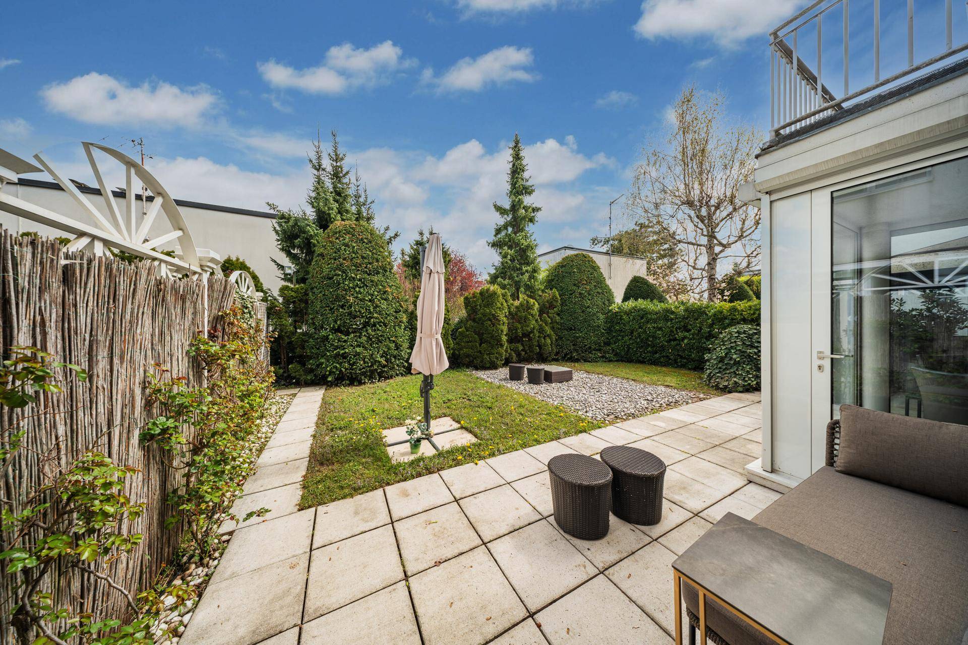 Gepflegter Gartenbereich mit Terrasse, Rasenfläche und üppiger Bepflanzung unter blauem Himmel.