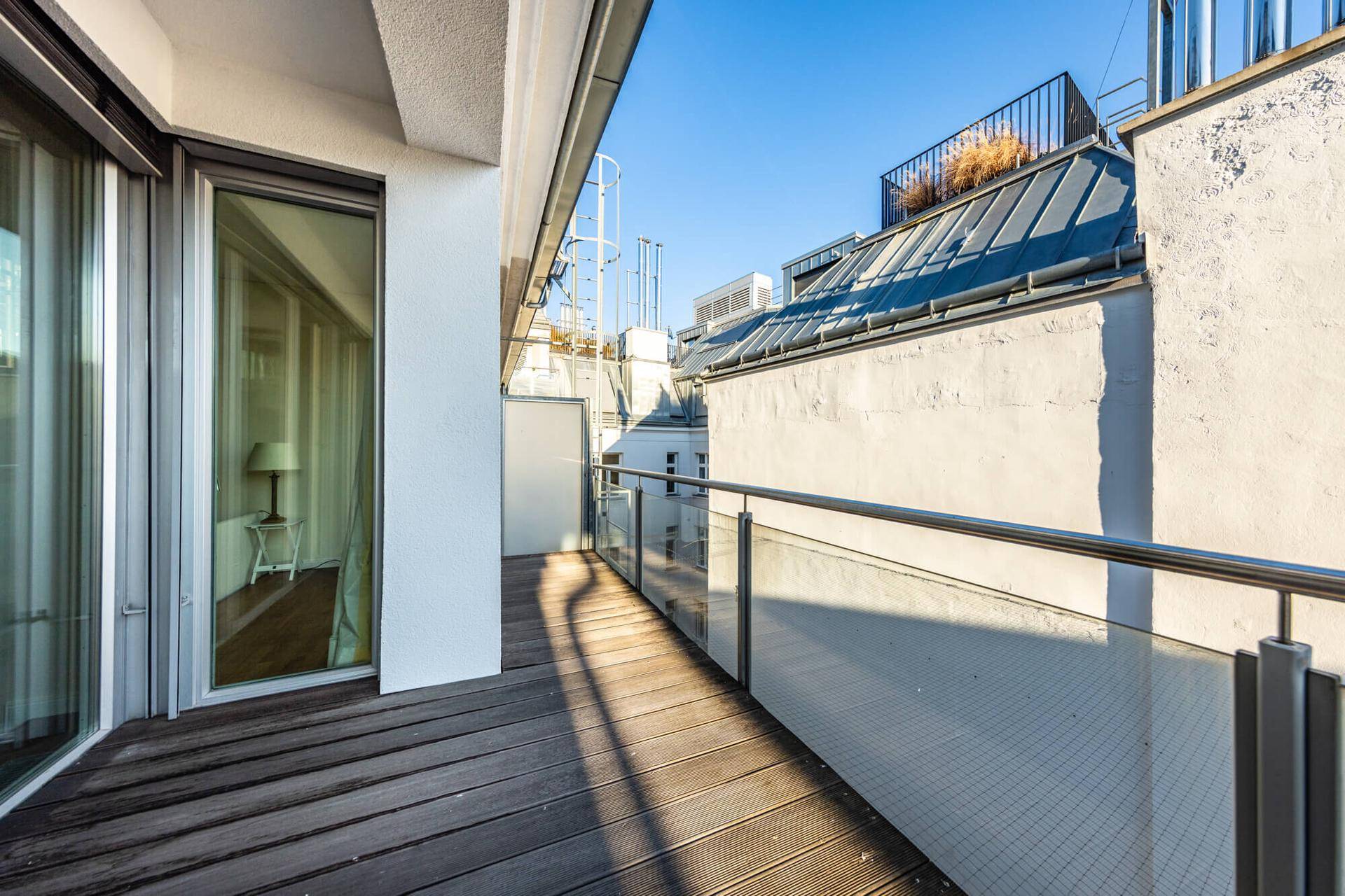 Langer Balkon mit Holzbelag und Glasgeländer, bietet einen Blick über die Dächer der Stadt.