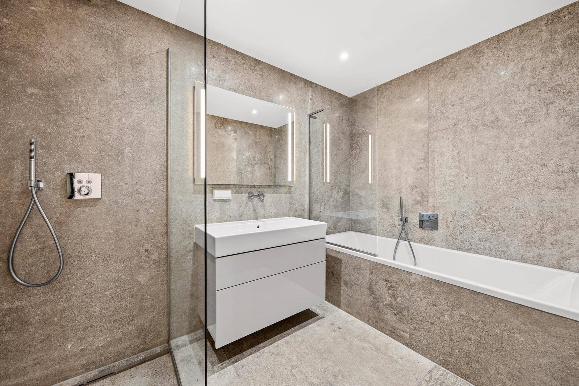 Modernes Badezimmer mit grauen Fliesen, Badewanne mit Glasabtrennung und elegantem Waschtisch.