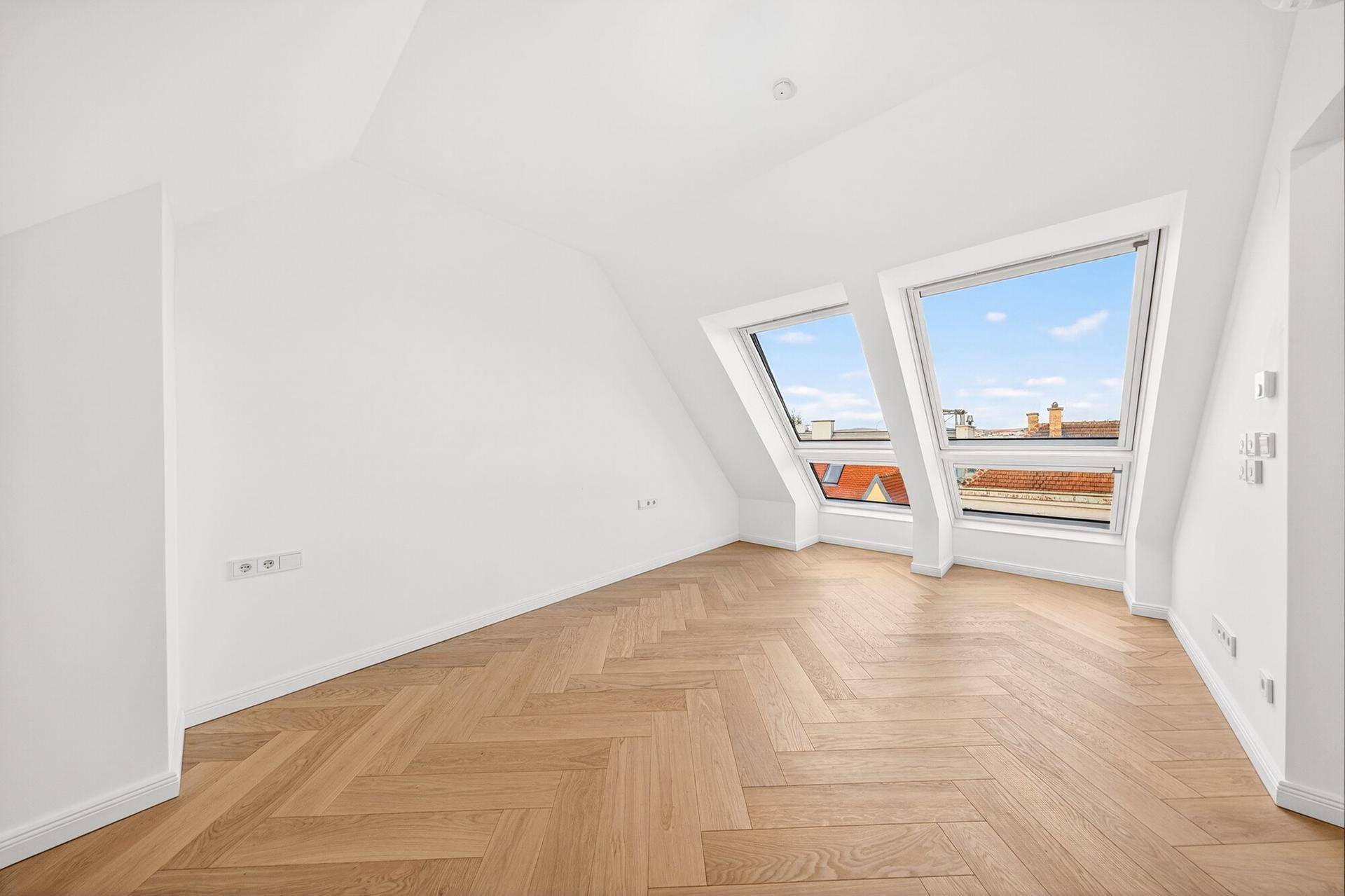 Heller, leerer Raum mit Fischgrätparkett und zwei großen Dachfenstern mit Stadtblick.