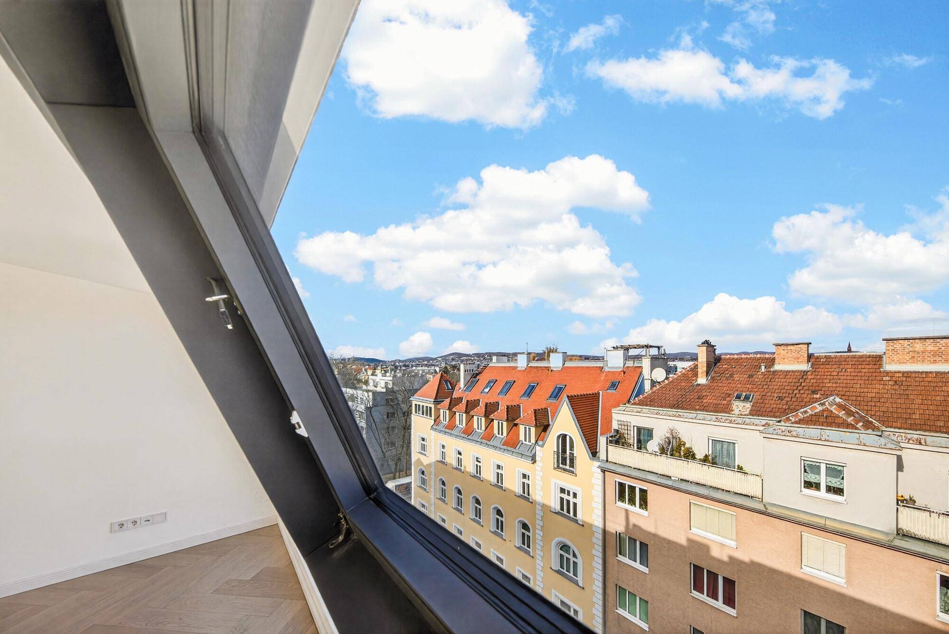 Blick aus einem Dachfenster auf die Dächer der umliegenden Gebäude und die Stadt in der Ferne.