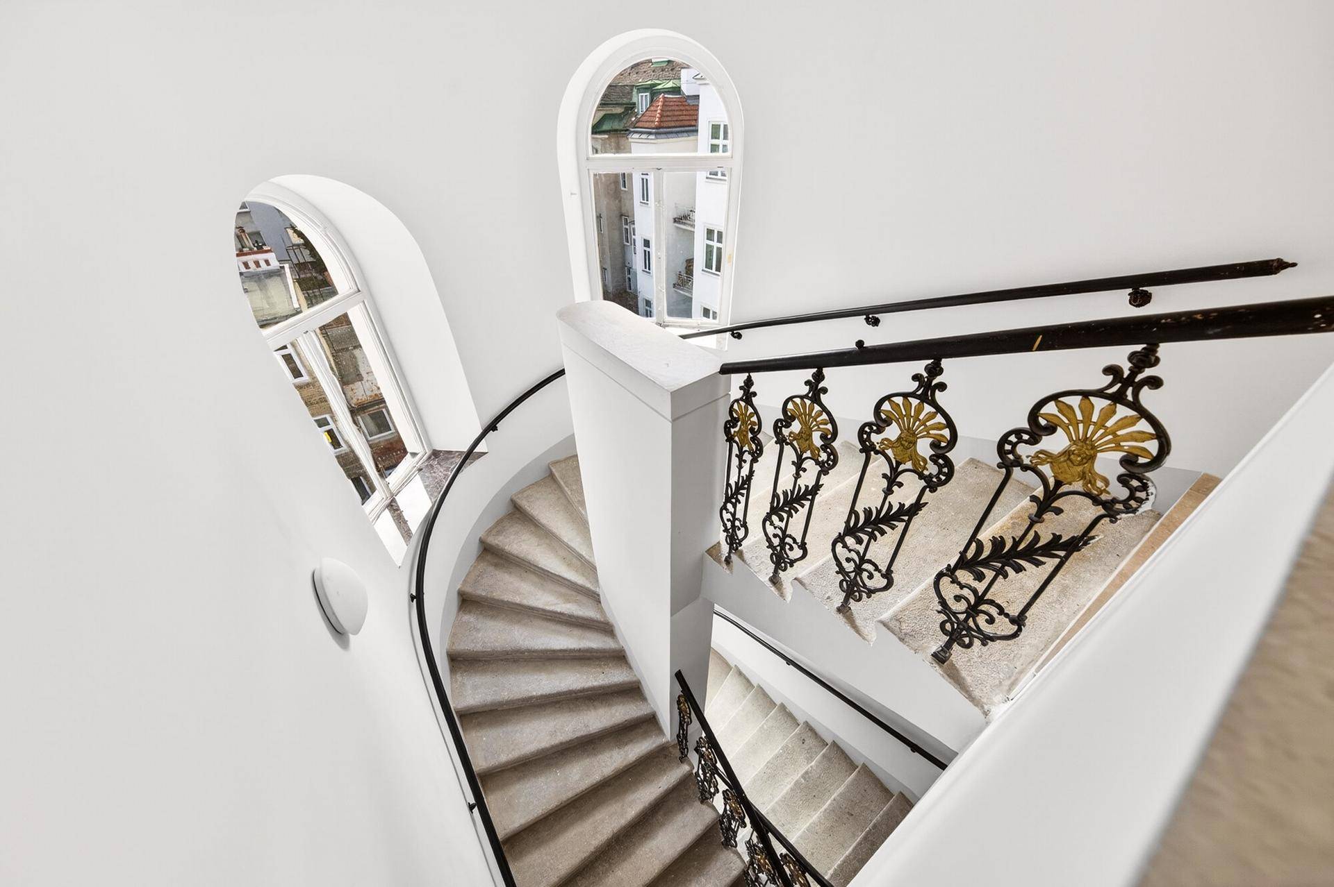 Elegantes, geschwungenes Treppenhaus mit kunstvollem Geländer und Teppichstufen, hell durch Bogenfenster.