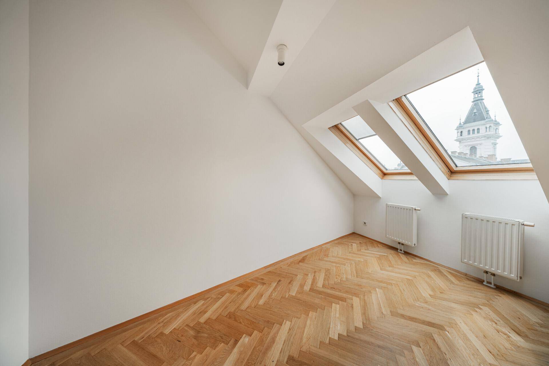 Gemütliches Zimmer mit Fischgrätparkett, Dachschräge und Dachfenstern mit Blick auf ein Gebäude.