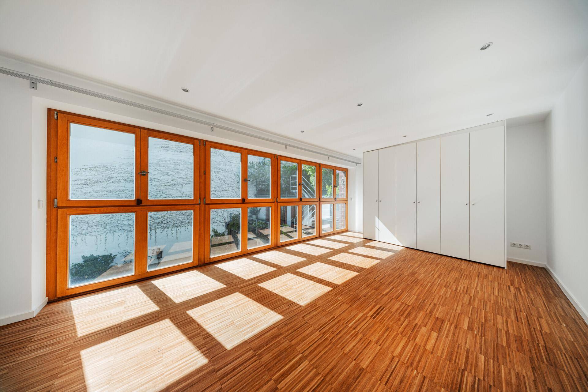 Schlafzimmer mit Einbauschränken, Holzparkett und großen Fenstern, die viel Licht hereinlassen.