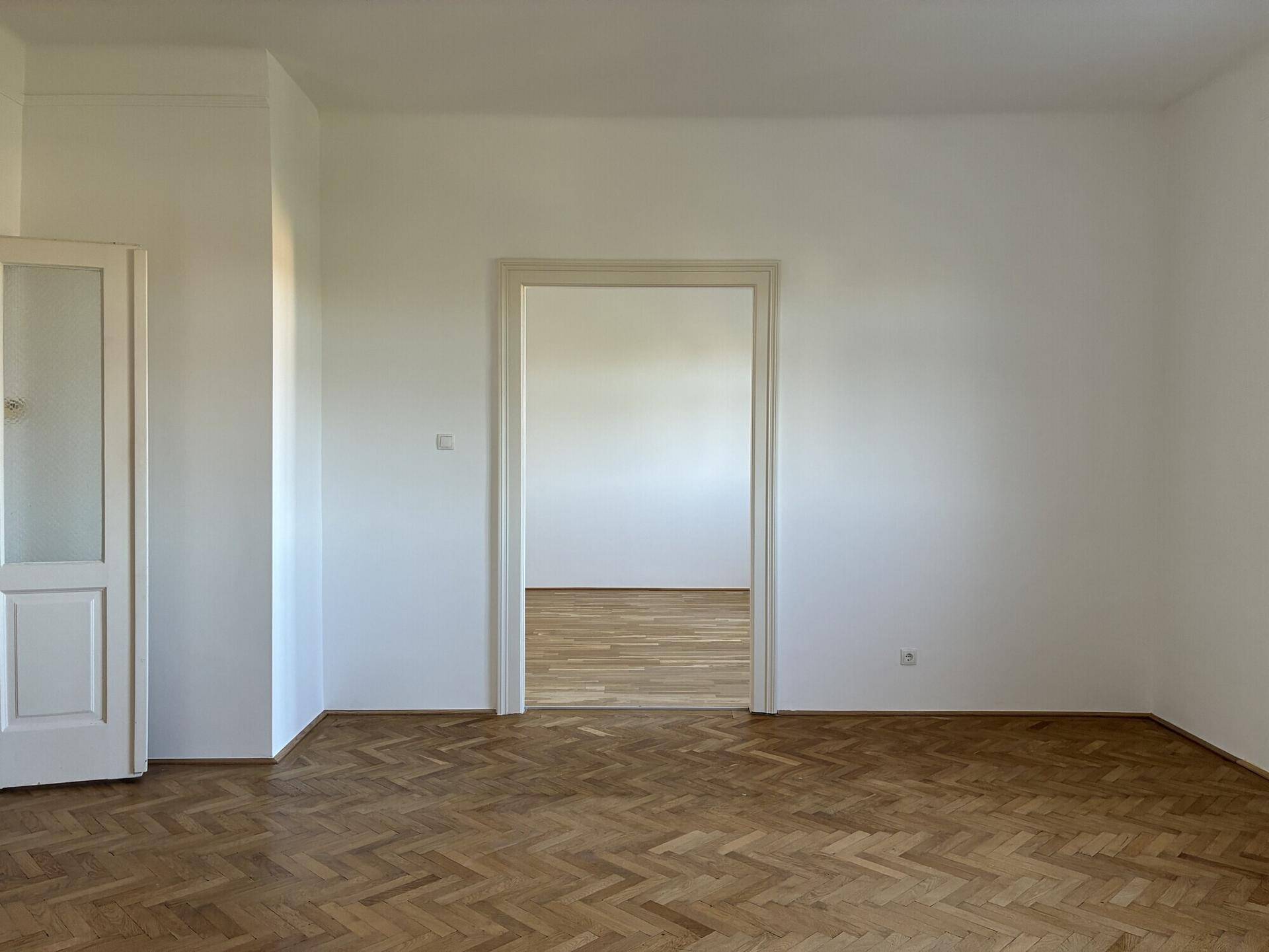 Helles, leeres Zimmer mit Fischgrätparkett und Durchgang zu weiteren Räumen.