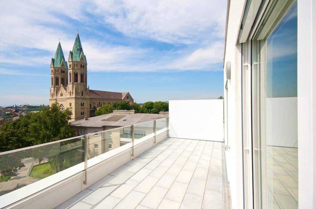 Weitläufige Dachterrasse mit Fliesenboden und Glasgeländer, bietet einen weiten Blick auf die Stadt und eine Kirche.