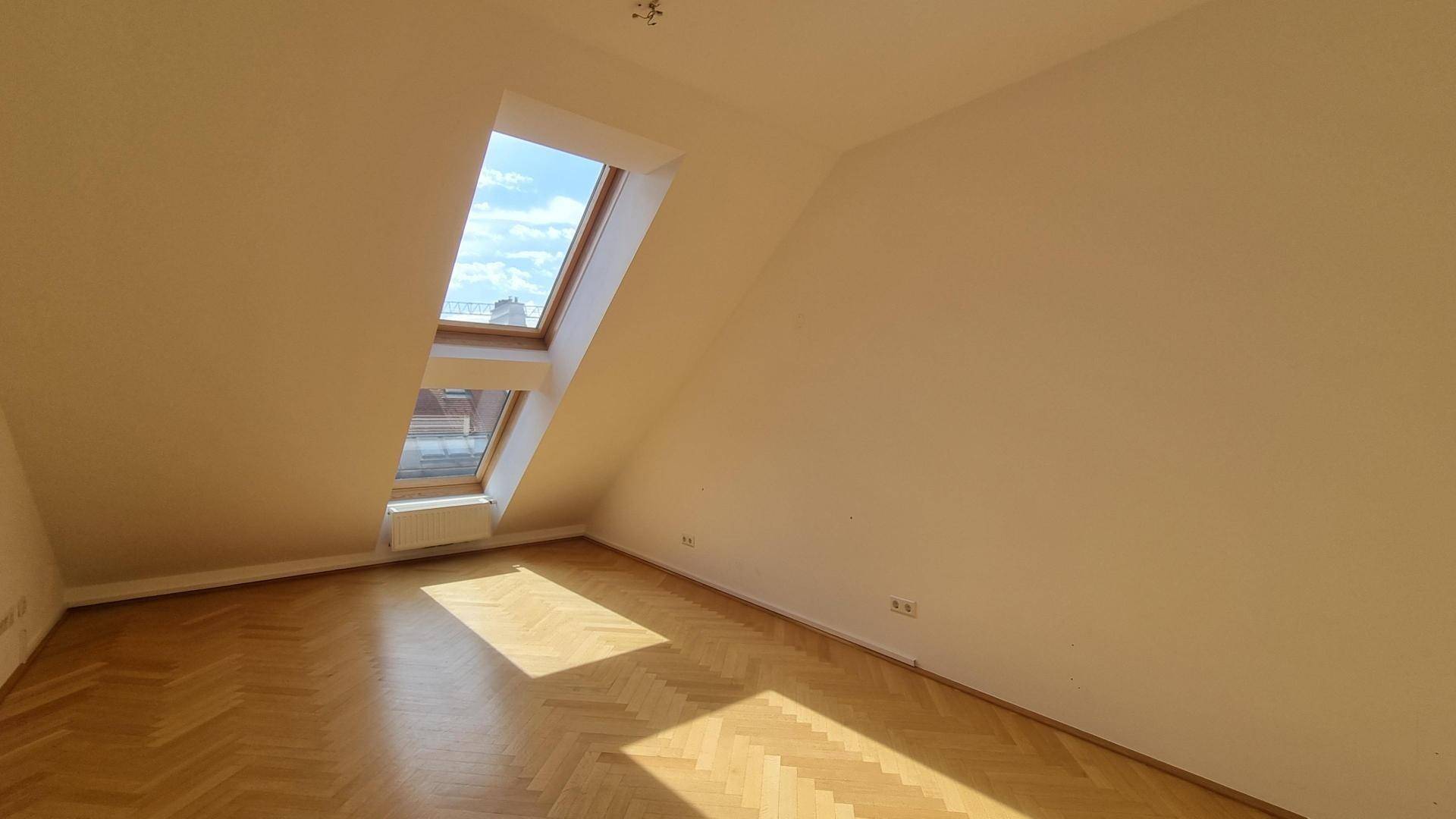 Helles Dachzimmer mit schrägen Wänden, einem Dachfenster und Fischgrätparkettboden.