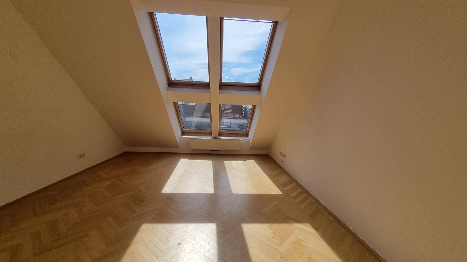 Helles Dachzimmer mit schrägen Wänden, zwei Dachfenstern und Fischgrätparkettboden.