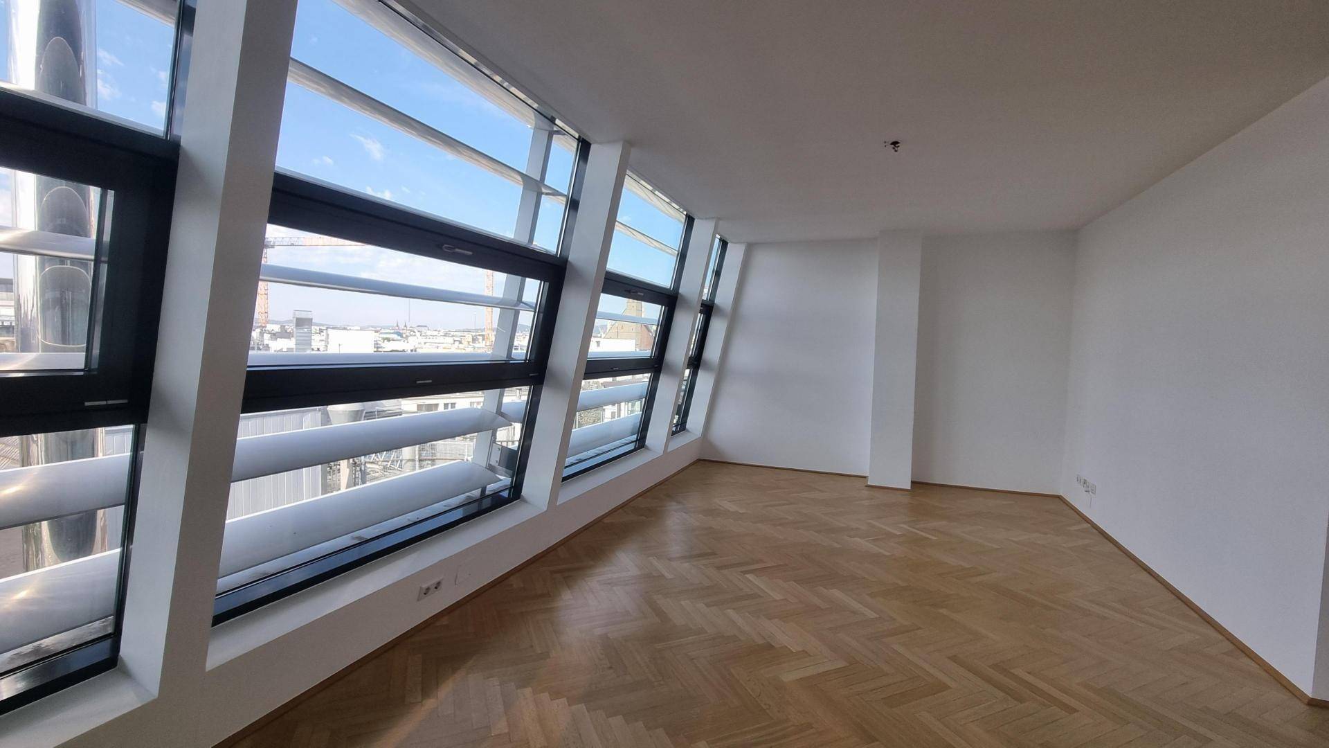 Geräumiger, leerer Wohnraum mit hellem Fischgrätparkett und schrägen Fensterfronten.