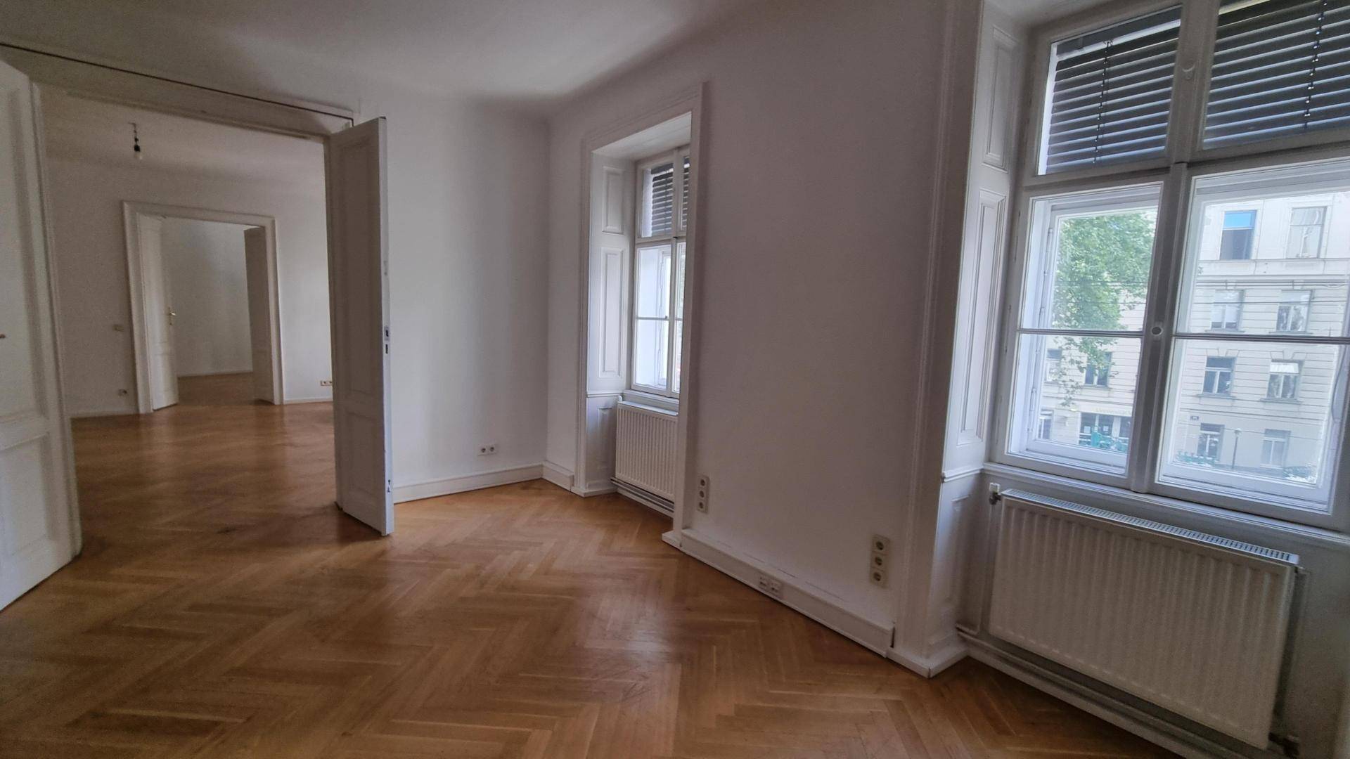 Geräumiges Zimmer mit Fischgrätparkett und hohen Fenstern, das in weitere Räume führt.