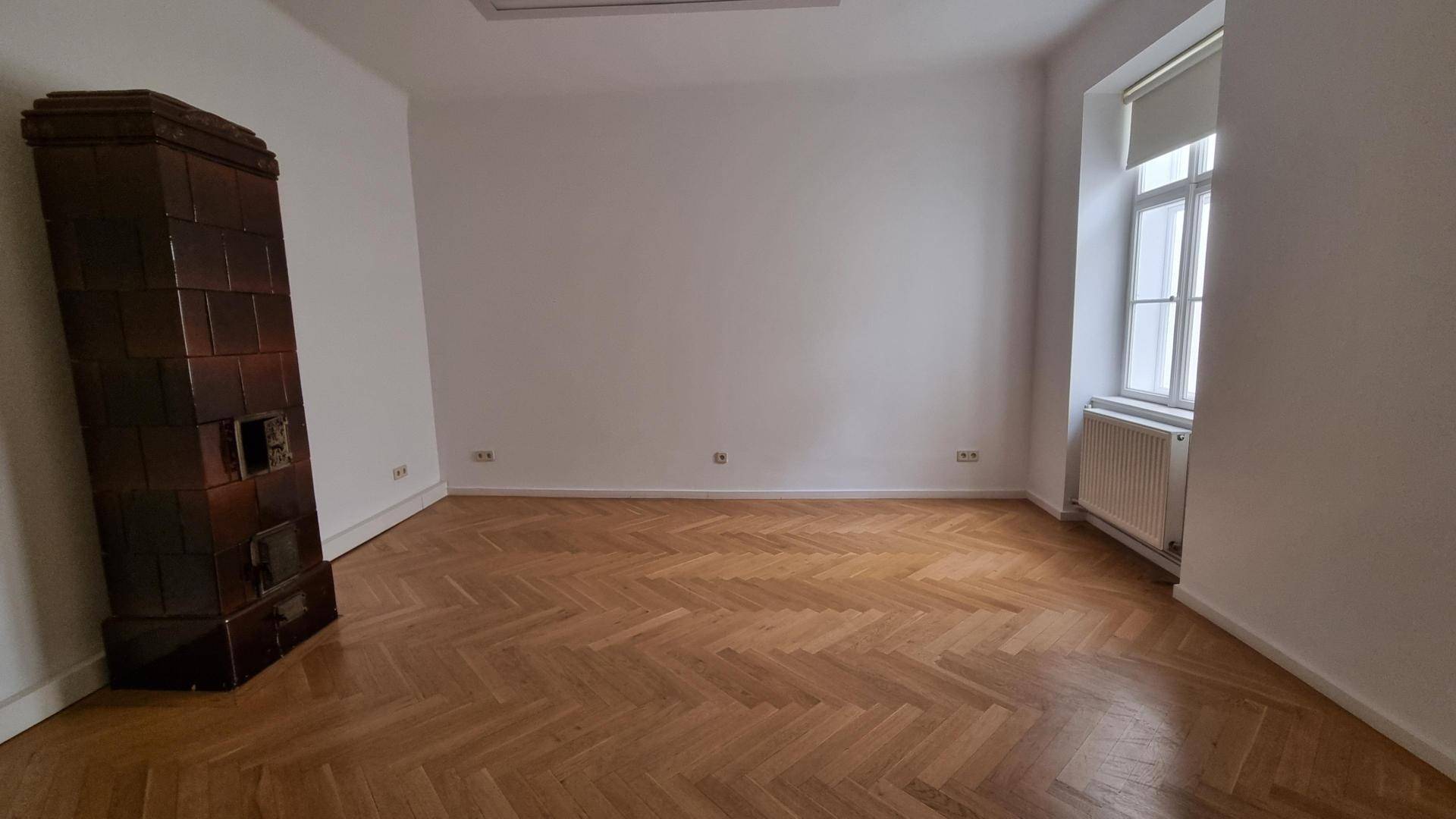 Helles Zimmer mit traditionellem Kachelofen und Fischgrätparkett, bereit zur individuellen Gestaltung.