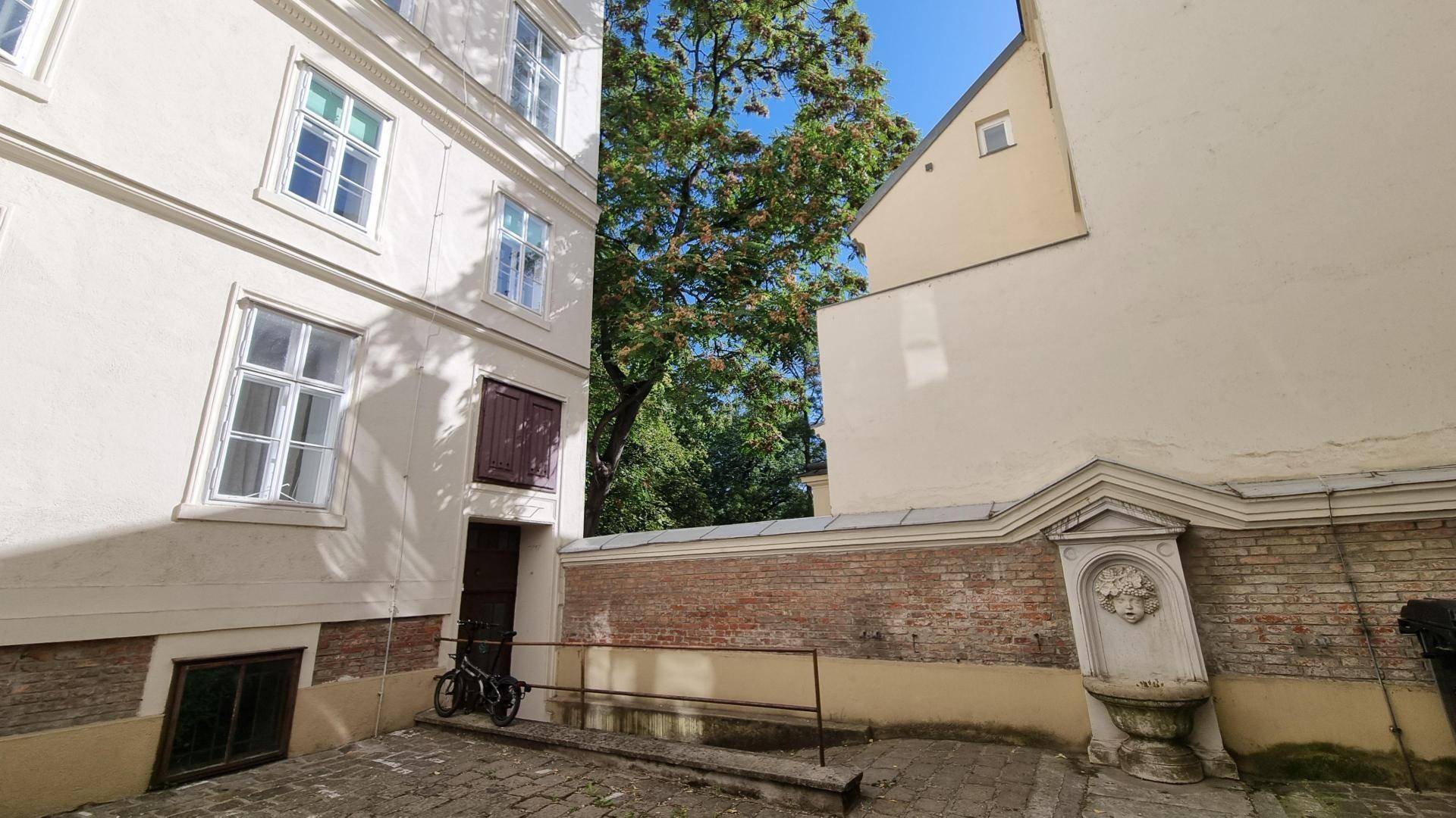 Ruhiger Innenhof mit altem Brunnen und grünen Bäumen, umgeben von Gebäudefassaden.