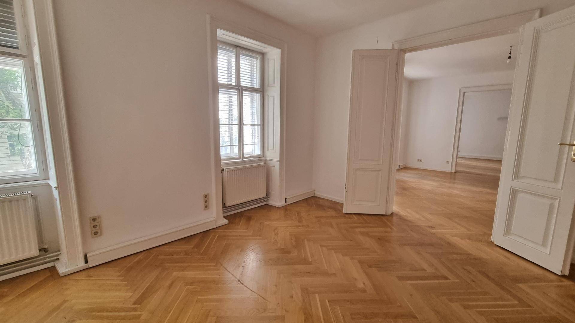 Helles Zimmer mit Fischgrätparkett und hohen Fenstern, das viel Tageslicht hereinlässt.