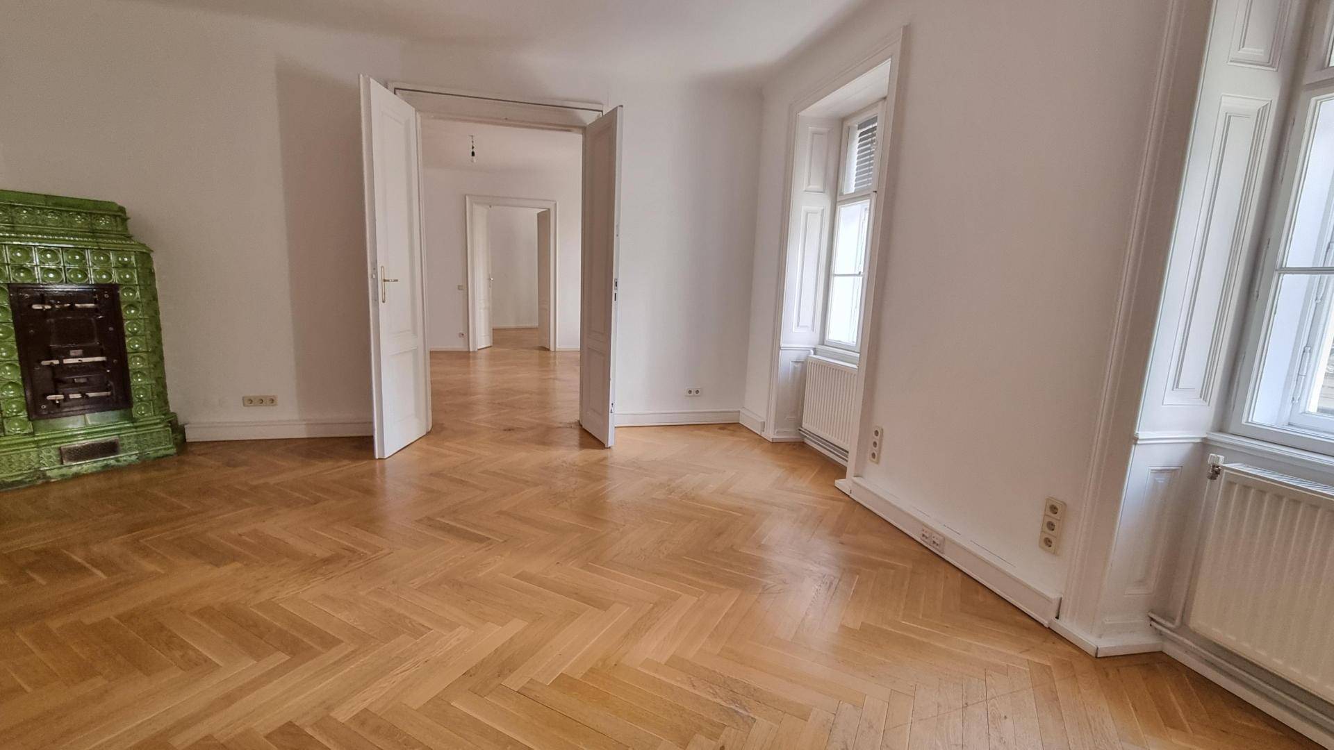 Geräumiges Zimmer mit traditionellem Kachelofen und Fischgrätparkett, das in weitere Räume führt.