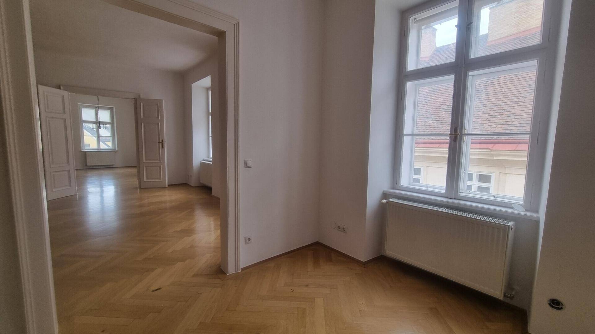 Helle Ecke eines Zimmers mit Fenster und Blick in einen weiteren Raum mit Parkettboden.