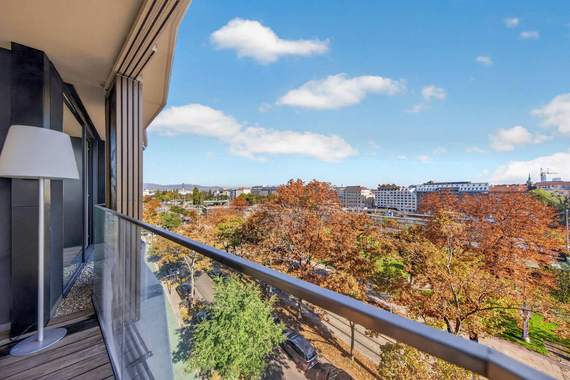 Balkon mit Glasbrüstung, Holzboden und weitem Blick über die Stadt und Bäume.