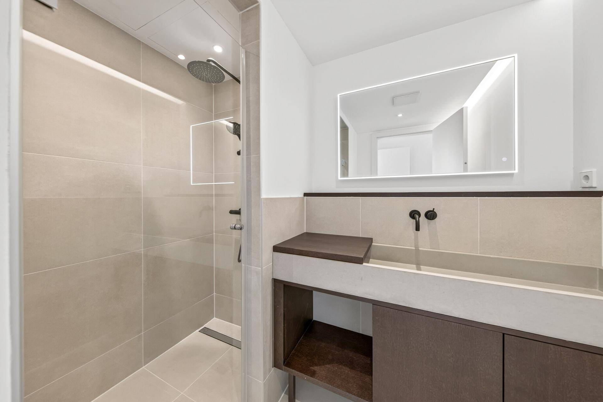 Modernes Badezimmer mit begehbarer Dusche, großformatigen Fliesen und beleuchtetem Spiegel.