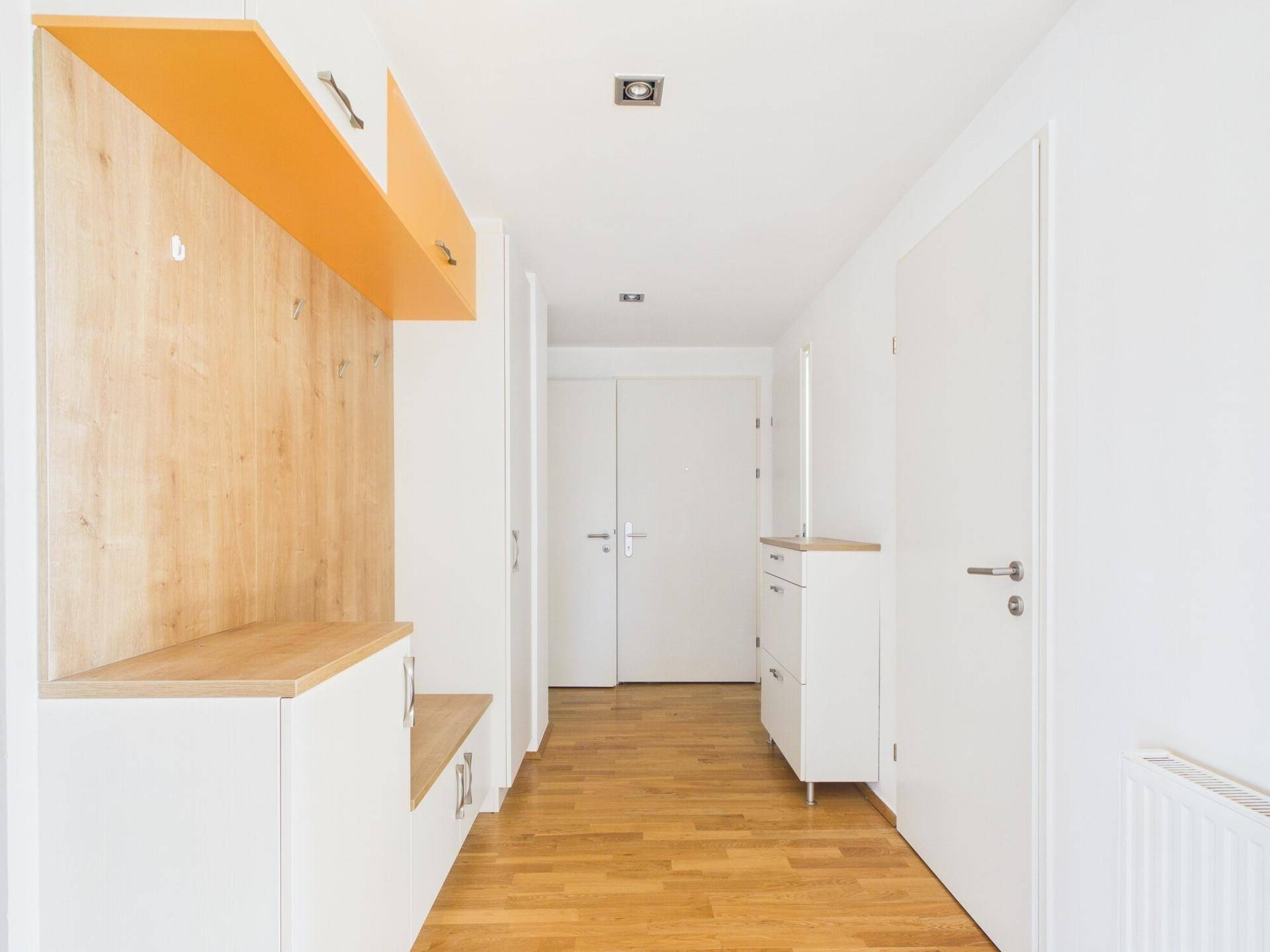 Langer Flur mit Holzboden, weißen Türen und integrierten Garderobenmöbeln in Holz und Orange.