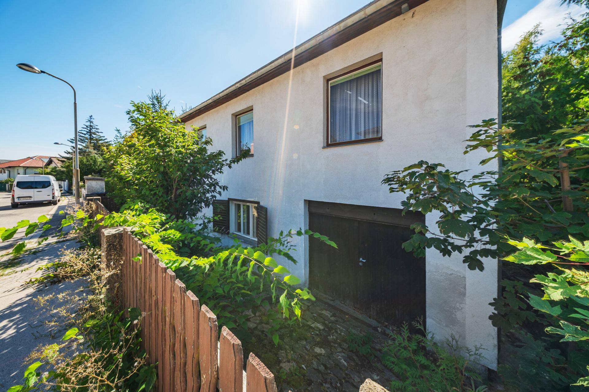 Seitenansicht des Hauses mit Garagentor, Fenstern und angrenzender Straße unter hellem Sonnenlicht.