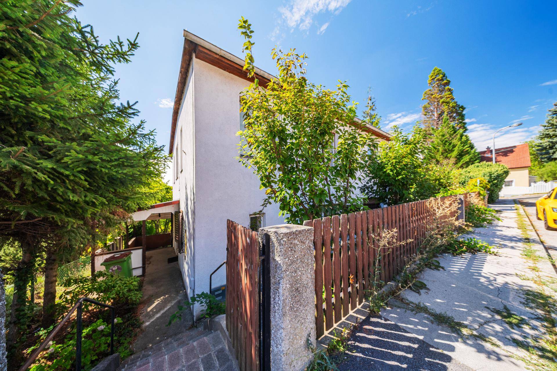Seitenansicht des Hauses mit Holzzaun und Zugang zum Grundstück von der sonnigen Straße.