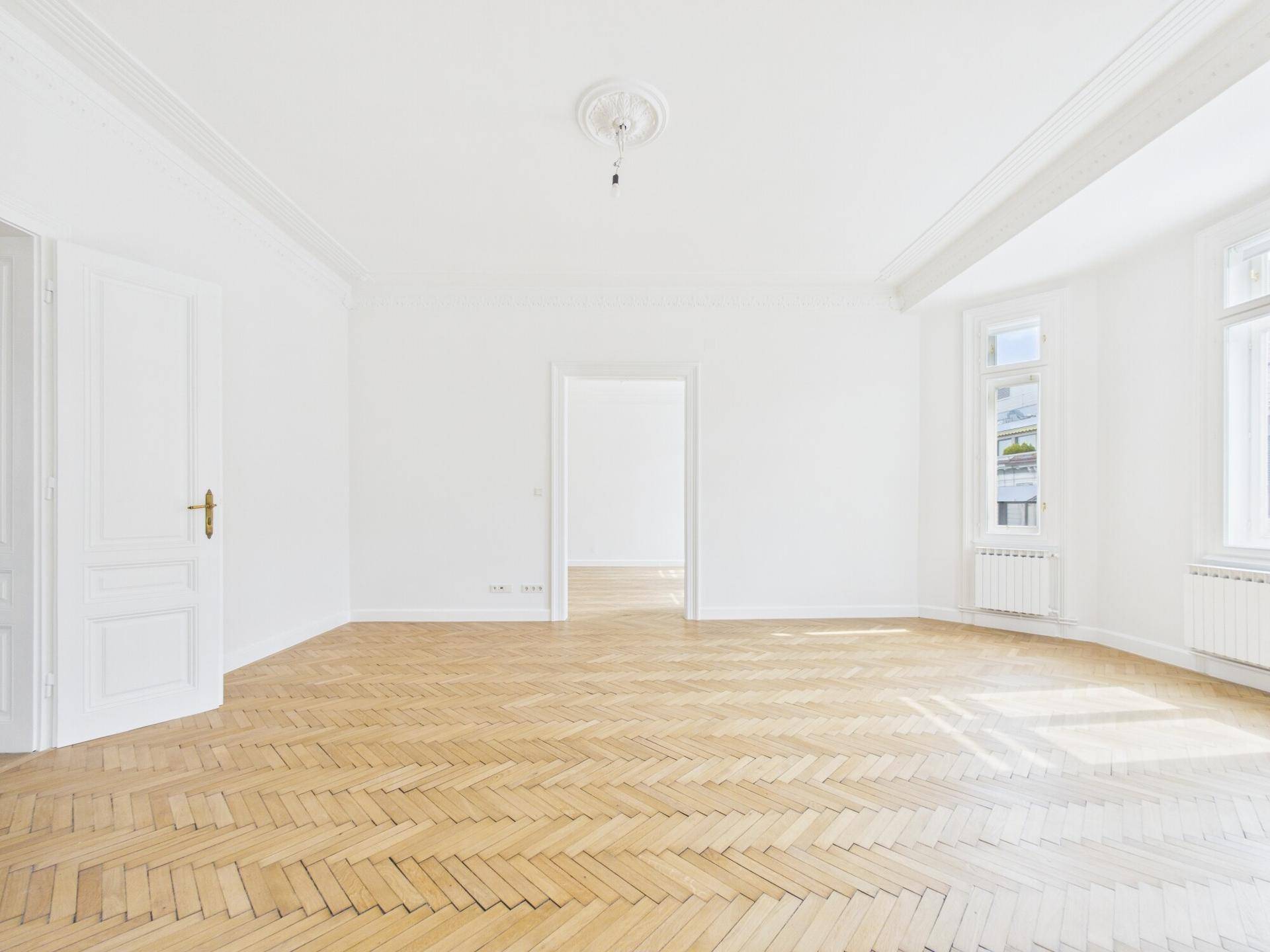 Geräumiges Zimmer mit Fischgrätparkett, hohen Decken und klassischer Tür mit Messinggriff.
