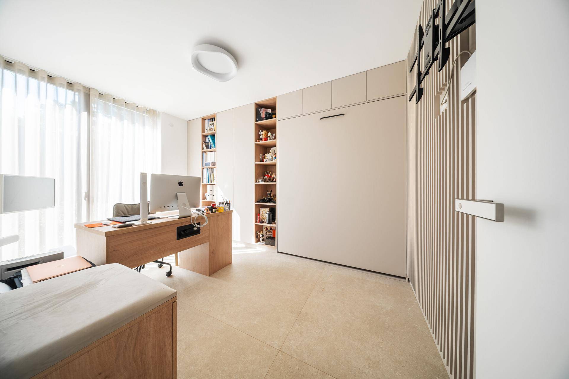 Helles Arbeitszimmer mit Schreibtisch, Einbauschränken und einem integrierten Klappbett für Gäste.