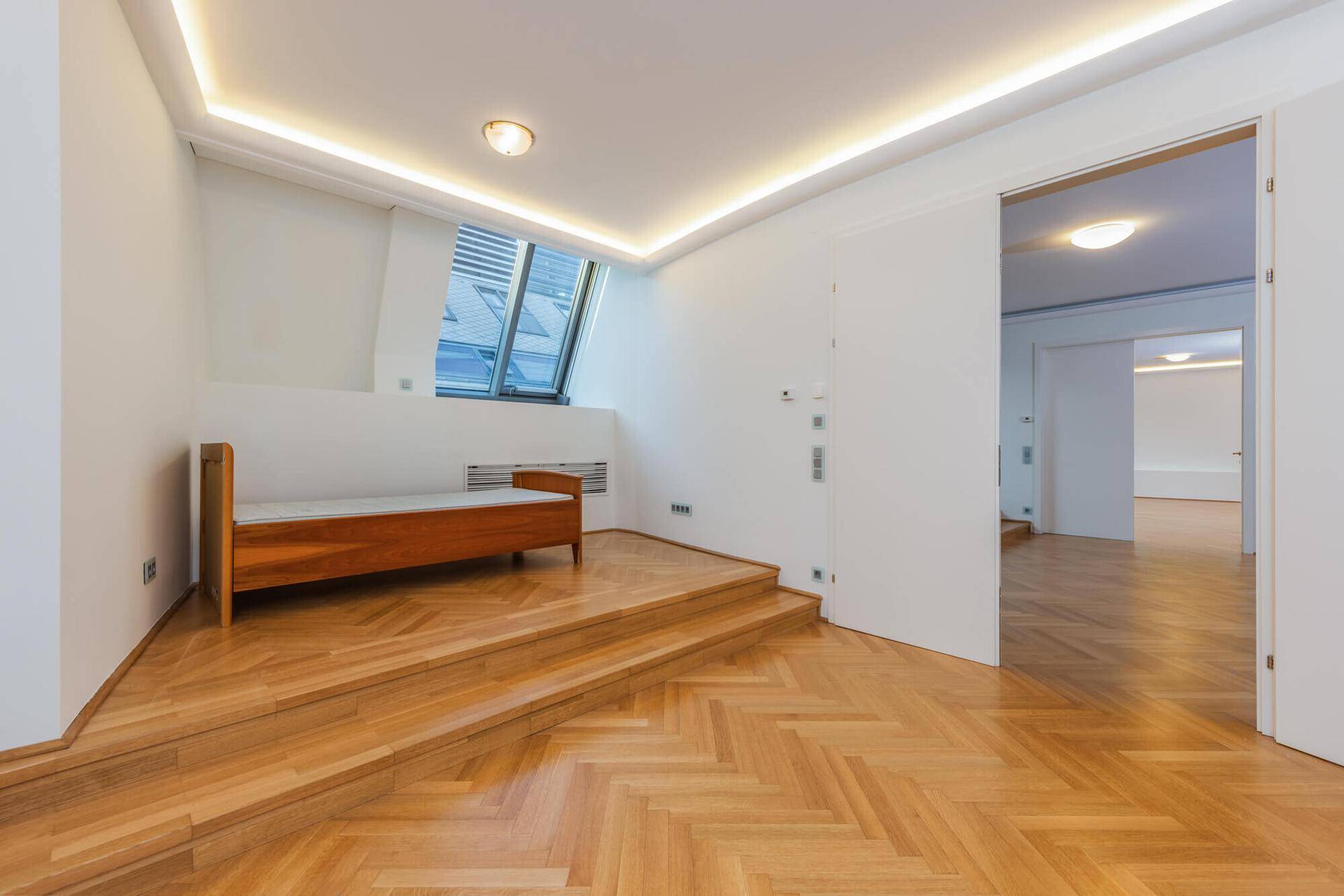 Schlafzimmer mit Holzparkett, Dachschrägenfenstern und indirekter Beleuchtung, offen zu einem weiteren Raum.