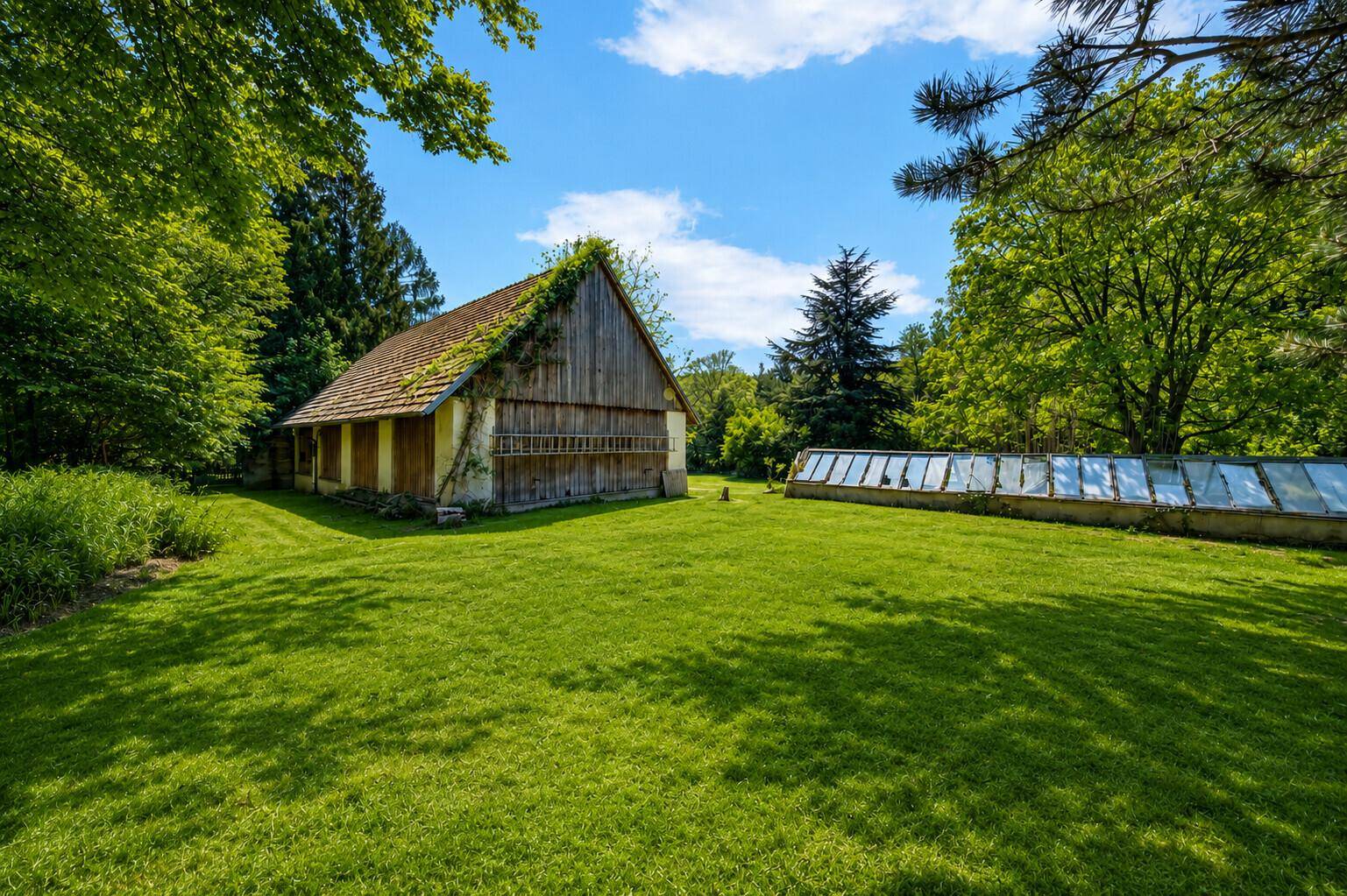 Rustikales Nebengebäude und Gewächshaus in einem weitläufigen grünen Garten unter blauem Himmel.