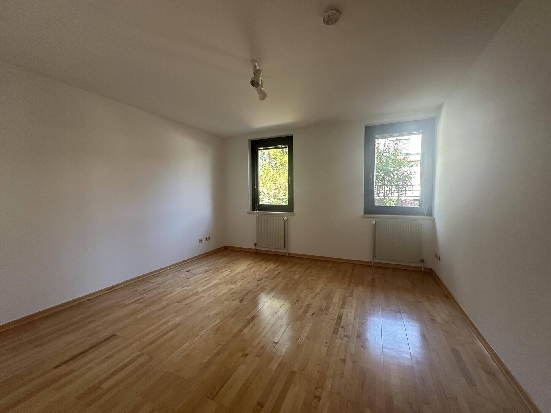 Helles Schlafzimmer mit Holzboden und zwei Fenstern, die viel Licht hereinlassen.