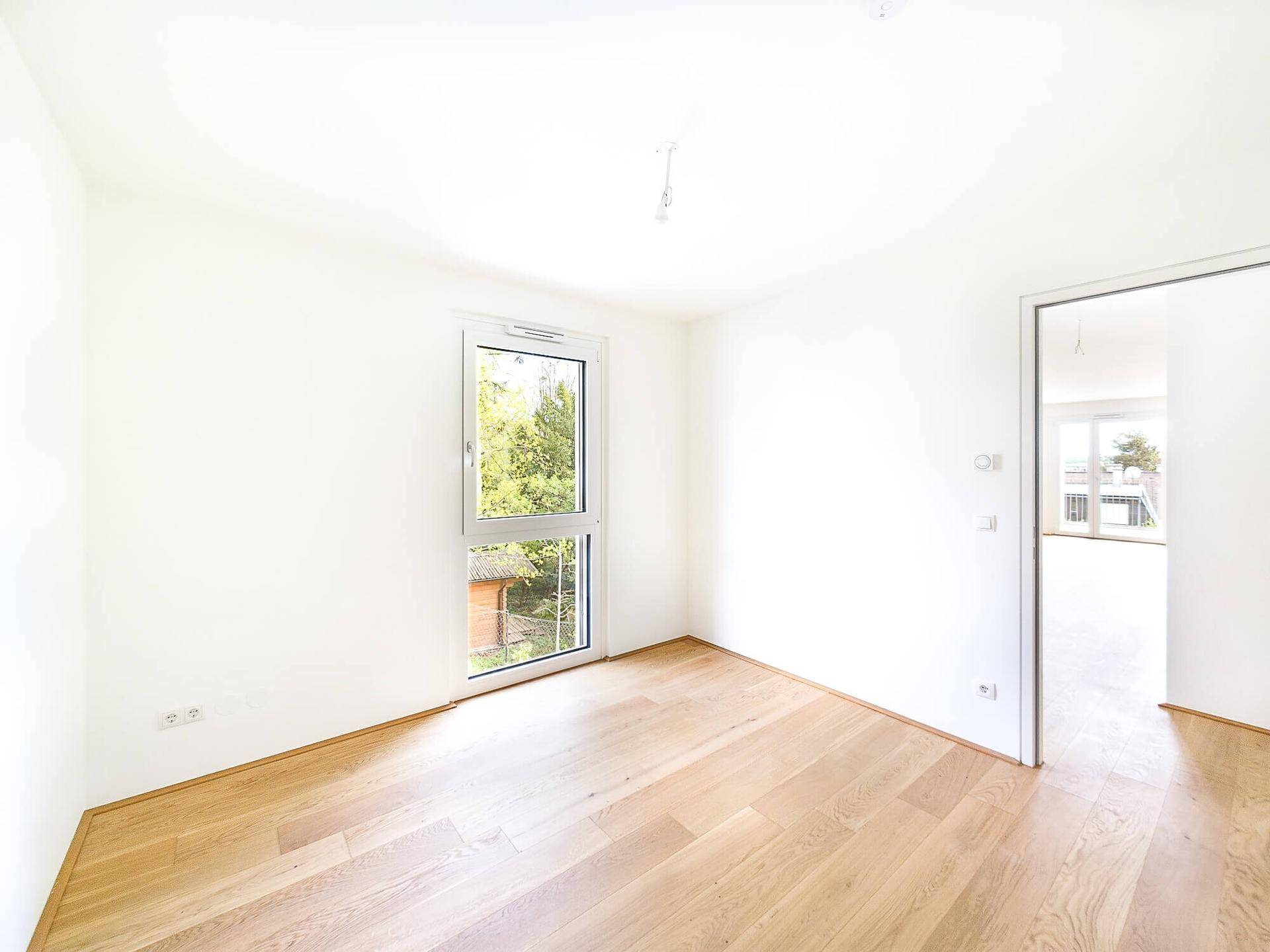 Helles Schlafzimmer mit Eichenparkett und großem Fenster mit Grünblick.