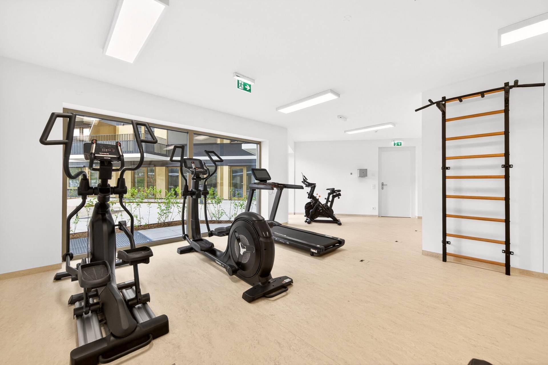 Heller Fitnessraum mit verschiedenen Trainingsgeräten und großen Fenstern für natürliches Licht.