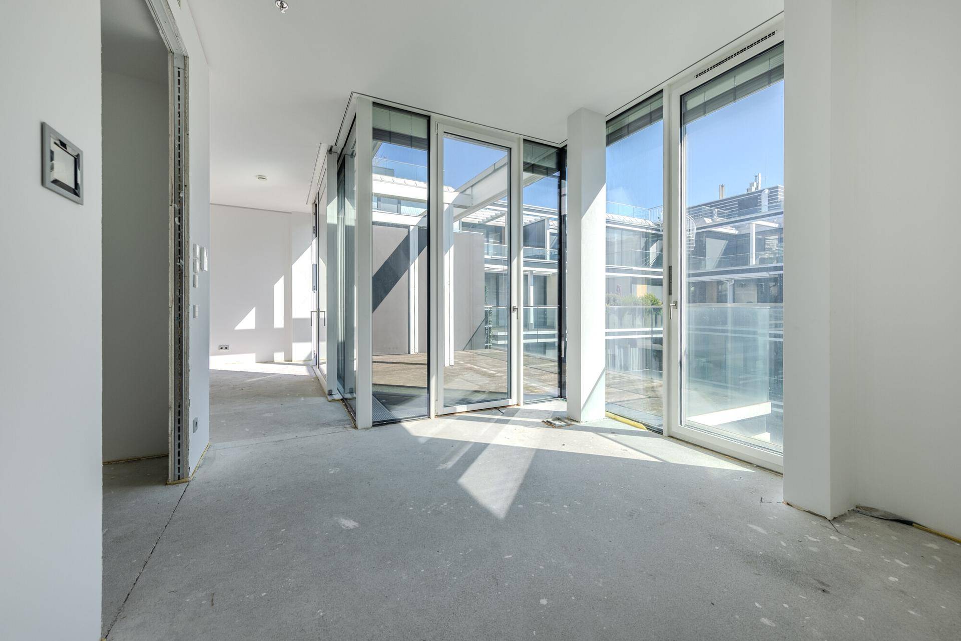 Unfertiger Innenraum mit Betonboden und großen Fenstern, bereit für den Ausbau und mit Blick auf den Innenhof.