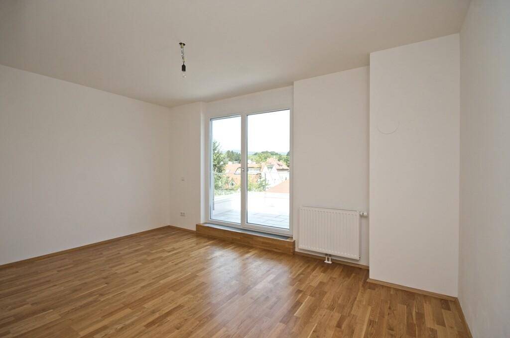 Geräumiges Zimmer mit Holzboden und großer Fenstertür, die viel Tageslicht hereinlässt und einen Ausblick bietet.