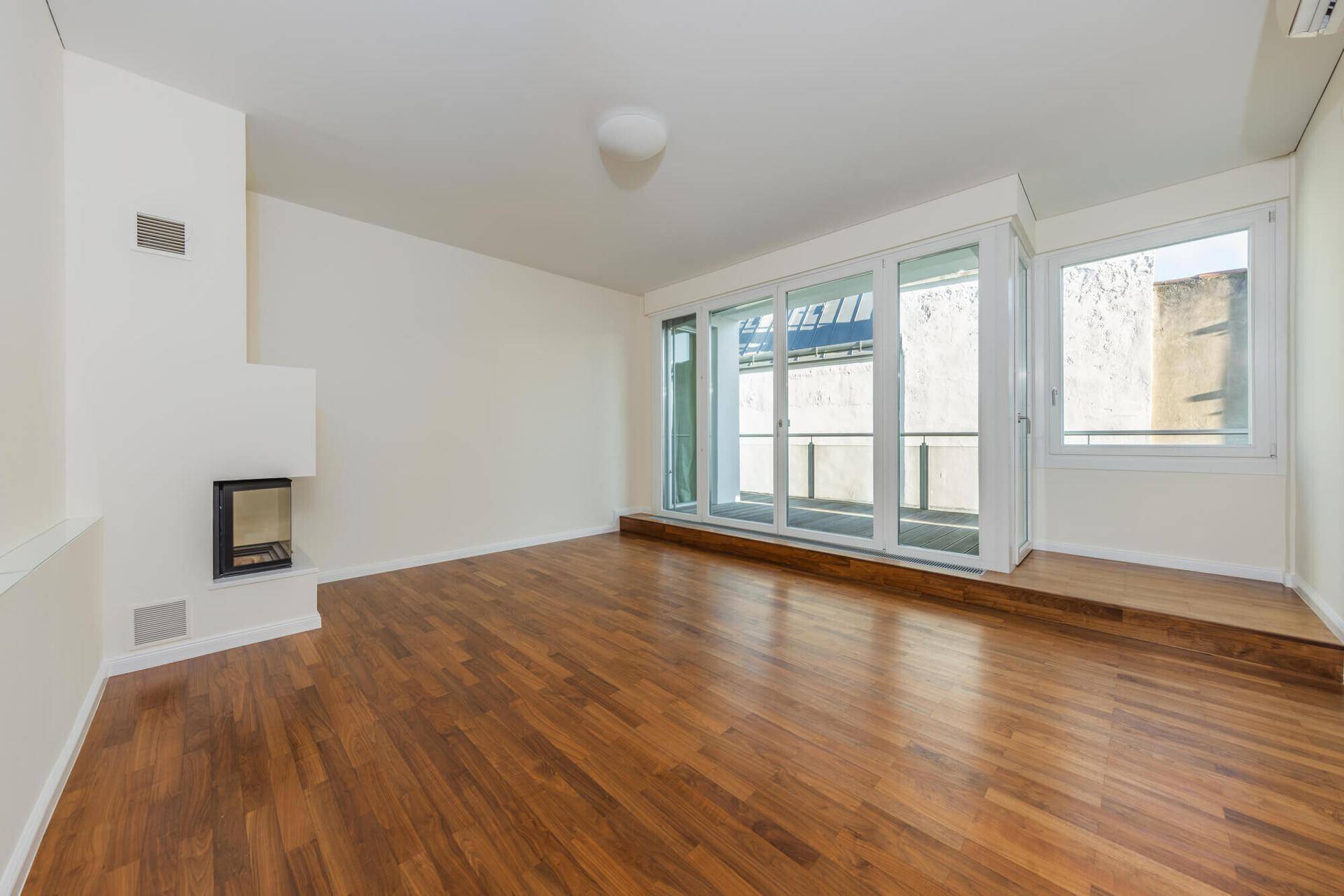 Unmöbliertes Wohnzimmer mit Kamin und großen Glasfronten, die viel Tageslicht hereinlassen.