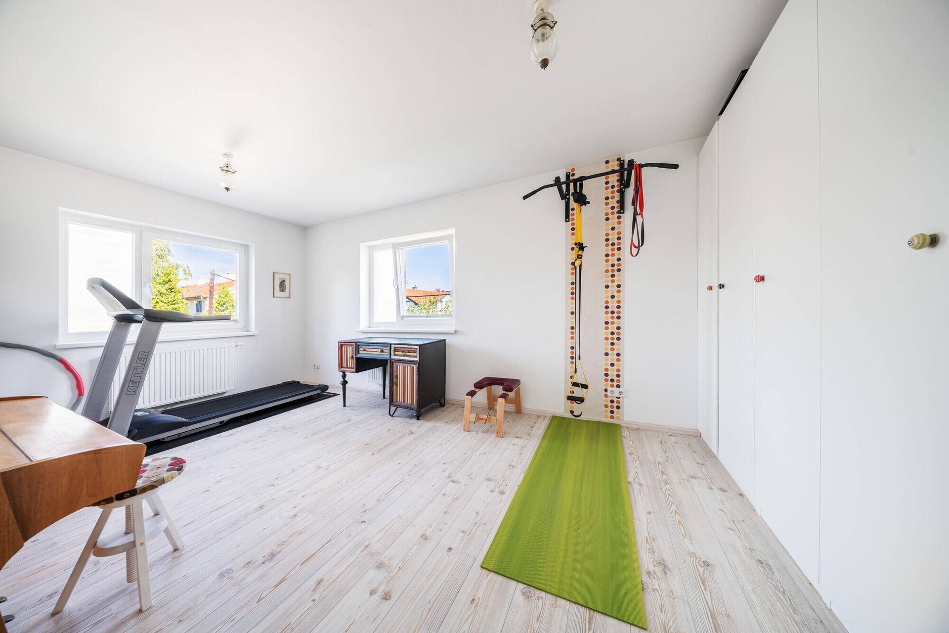 Heller Raum mit Holzboden, Fitnessgeräten und einem Schreibtisch am Fenster.
