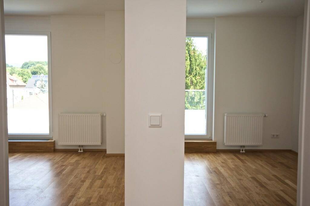 Heller, leerer Raum mit Holzboden, zwei Fenstern und Heizkörpern unter den Fensterbänken.