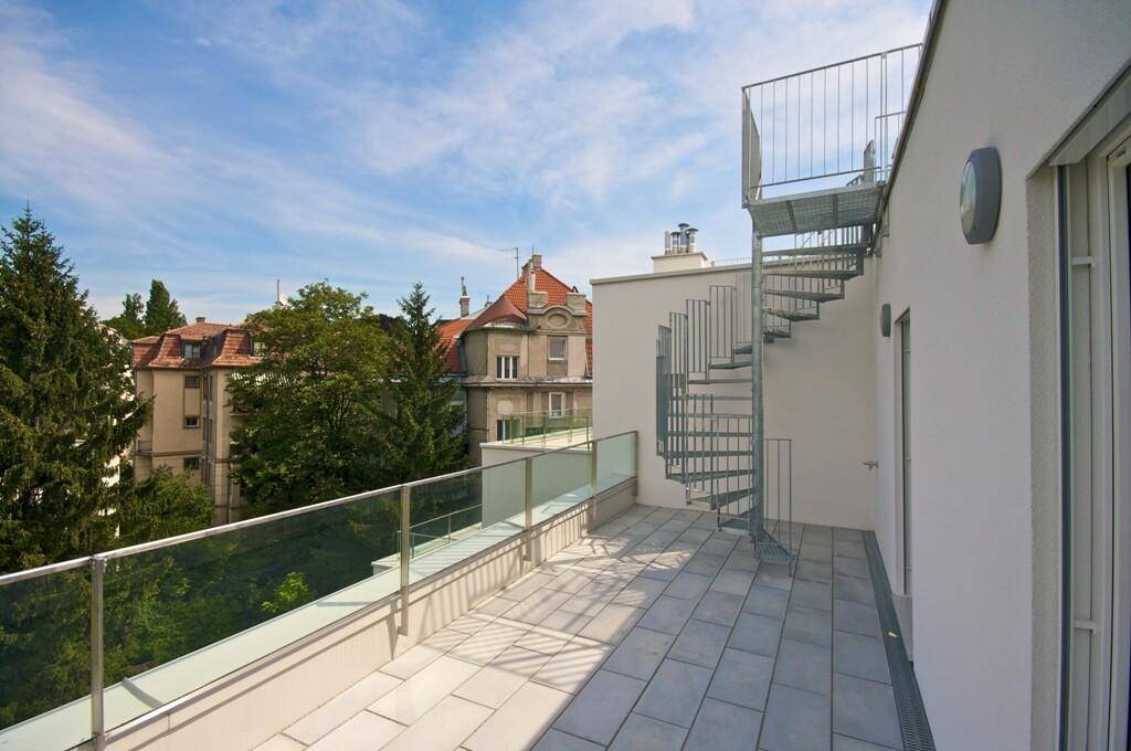 Große Dachterrasse mit Metallwendeltreppe, Glasgeländer und Blick auf die umliegenden Gebäude und Bäume.