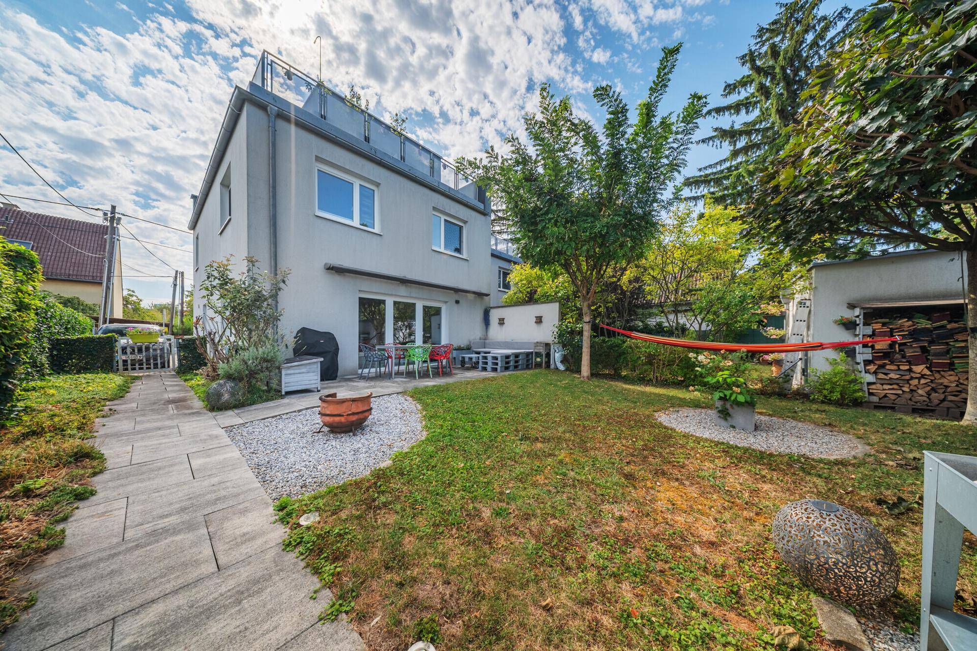 Rückseite des Hauses mit Garten, Terrasse, Rasenfläche und hohen Bäumen.