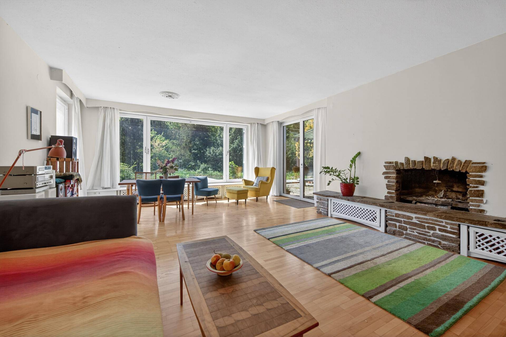 Helles Wohnzimmer mit Holzboden, großem Kamin und Blick ins Grüne durch Panoramafenster.