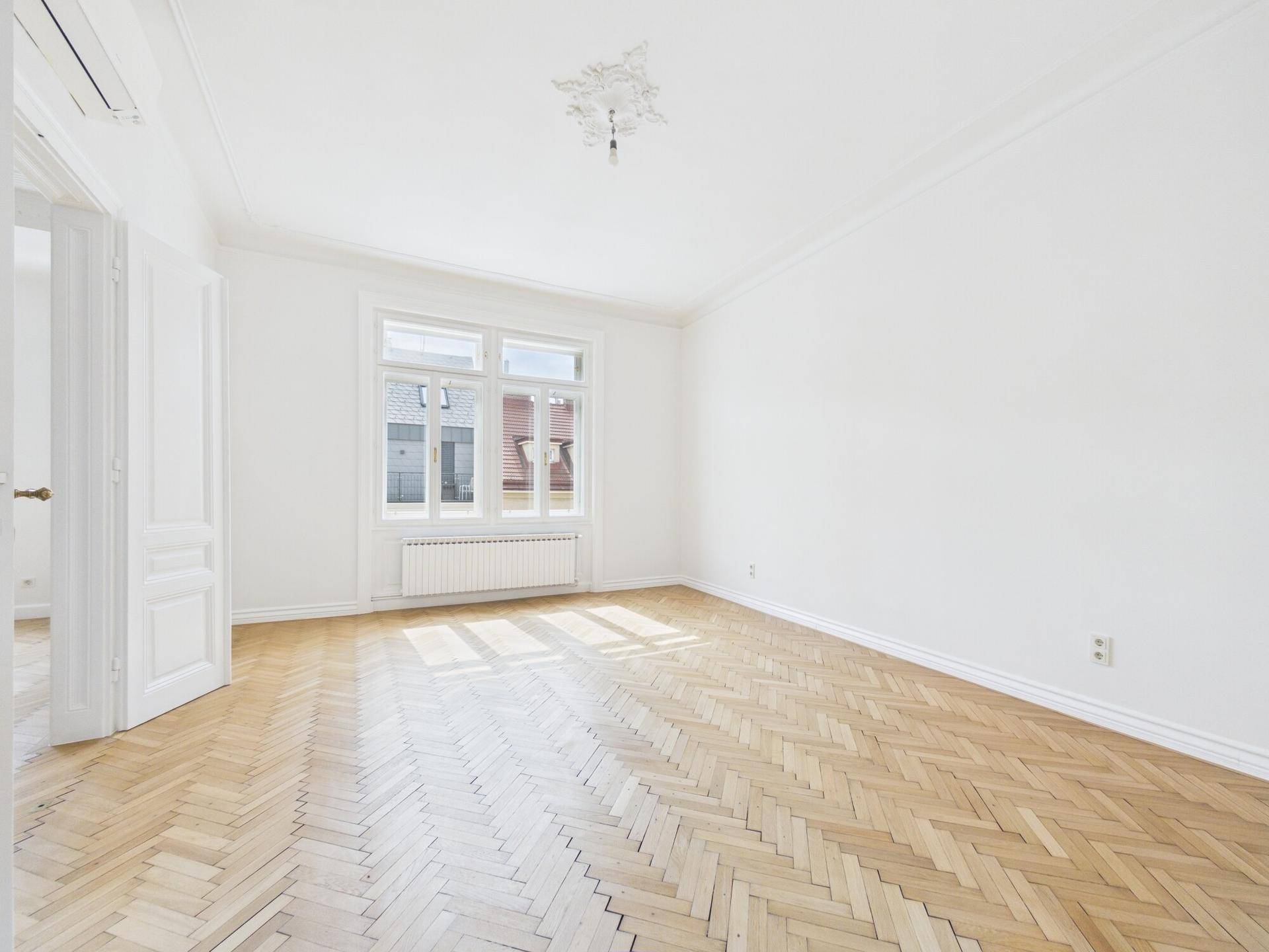 Helles Zimmer mit Fischgrätparkett, großem Fenster und Stuckverzierung an der Decke.