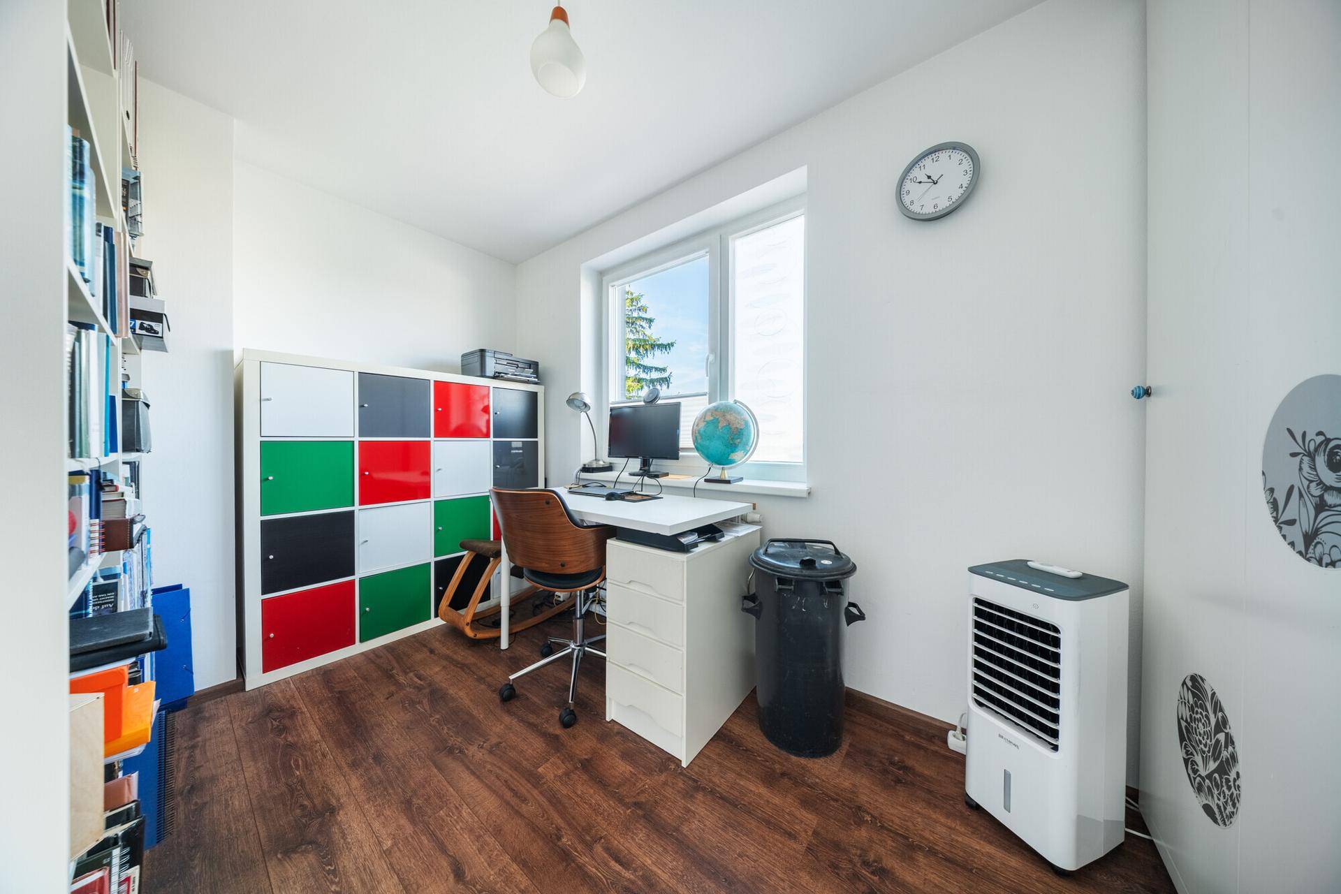 Arbeitszimmer mit Schreibtisch, Bücherregal und farbigen Schränken am Fenster.