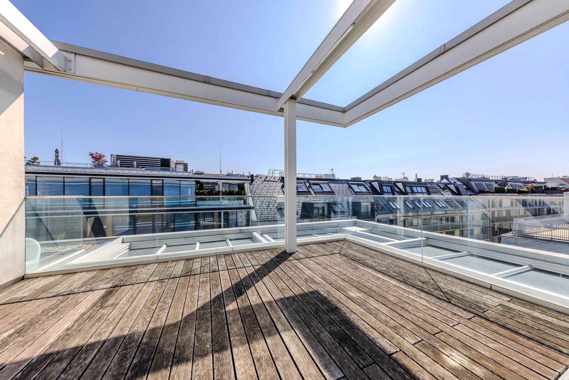 Sonnige Dachterrasse mit Holzdielen und modernem Glasgeländer, umgeben von Stadtgebäuden.