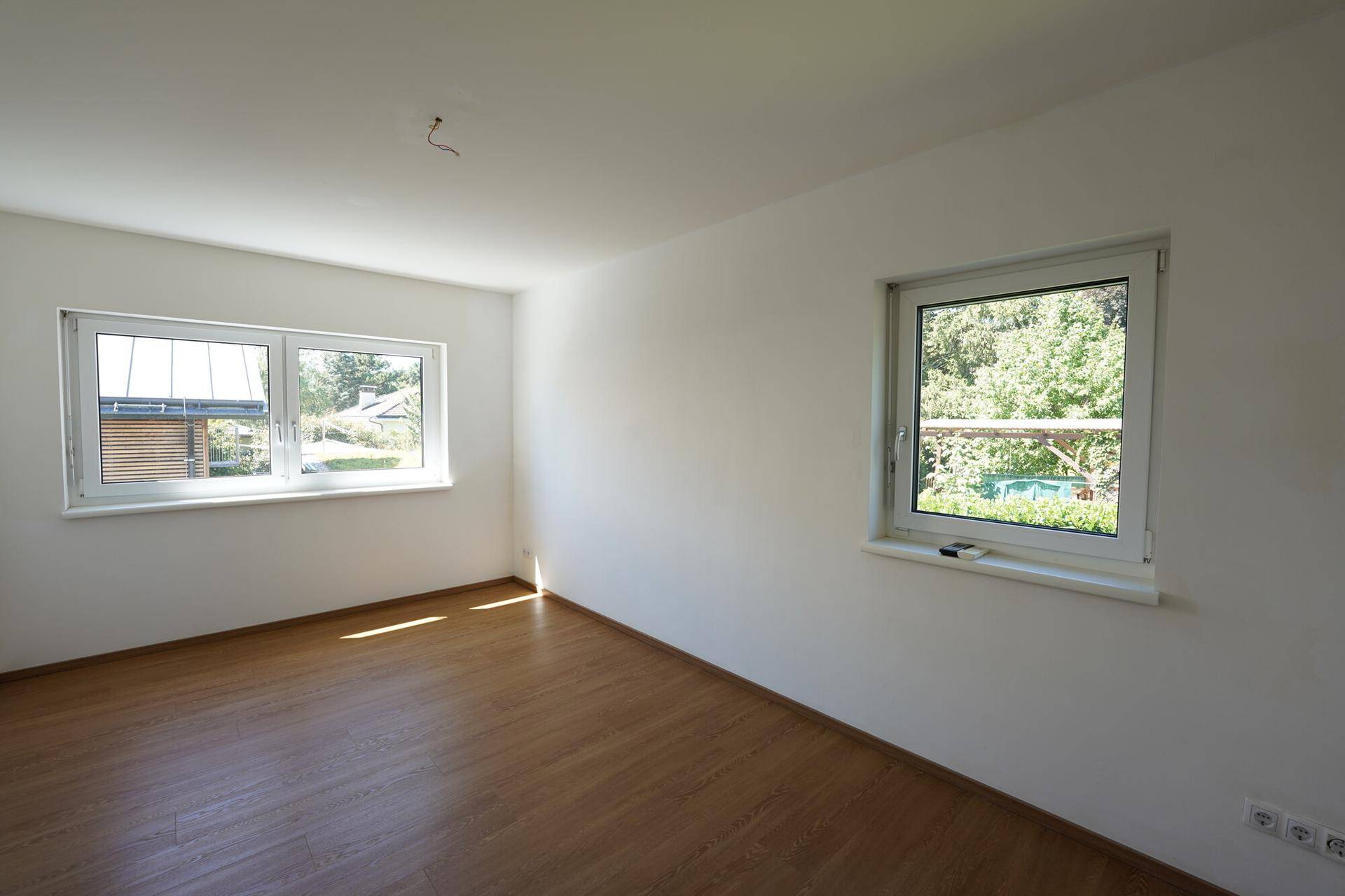 Helles, leeres Zimmer mit Laminatboden und zwei Fenstern, bereit zur individuellen Gestaltung.