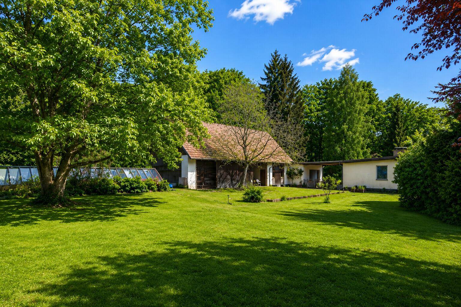 Weitläufiger Garten mit alten Bäumen, Nebengebäuden und einem Gewächshaus unter blauem Himmel.