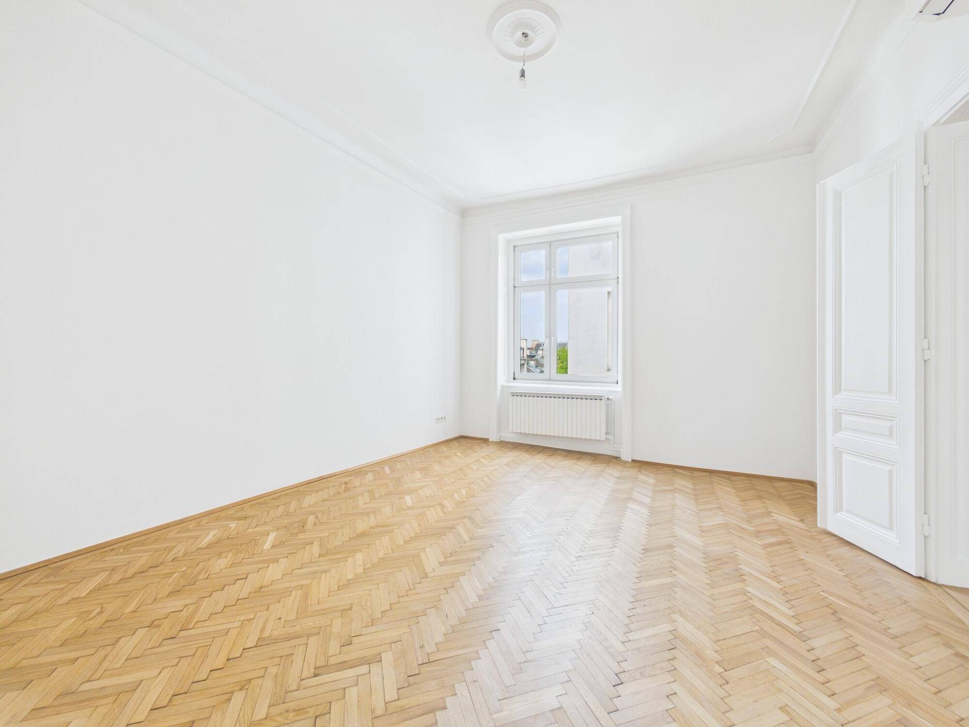 Leeres Zimmer mit Fischgrätparkett, einem Fenster und einer klassischen Flügeltür.