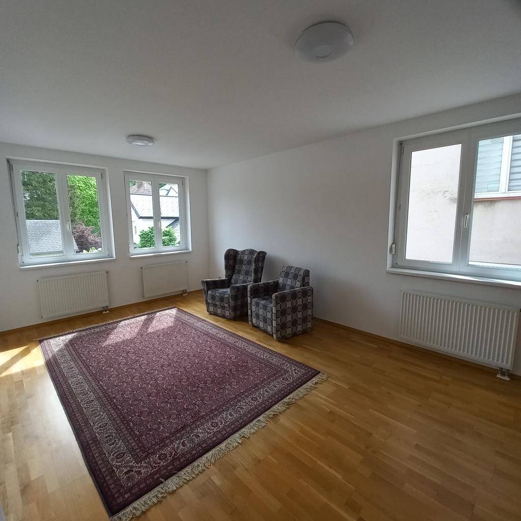 Geräumiges Wohnzimmer mit Holzfußboden, Teppich und viel Tageslicht durch die großen Fenster.